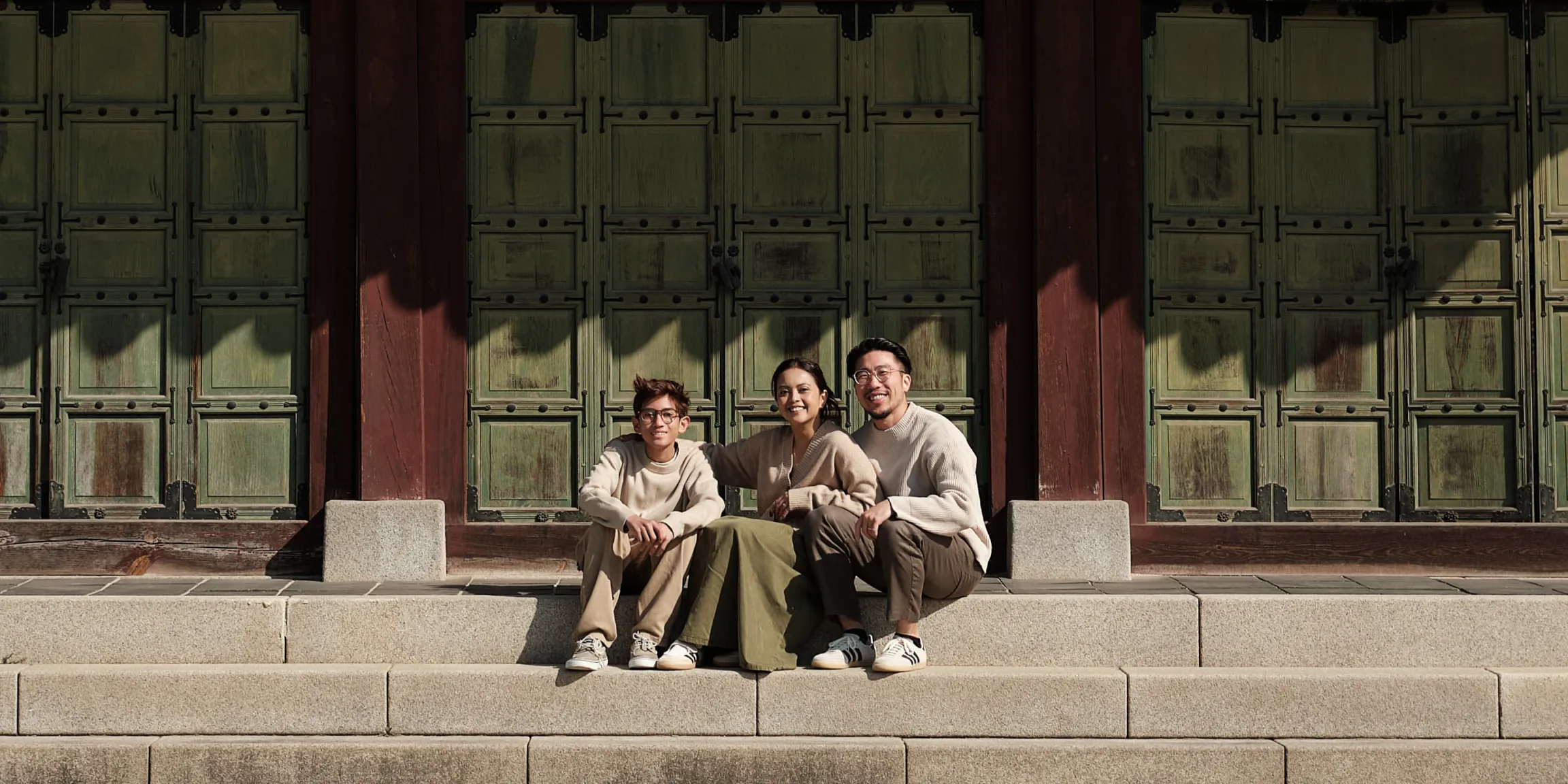 Phil Rhie with his wife and son, sitting close together on sunlit stone steps in front of tall green wooden doors